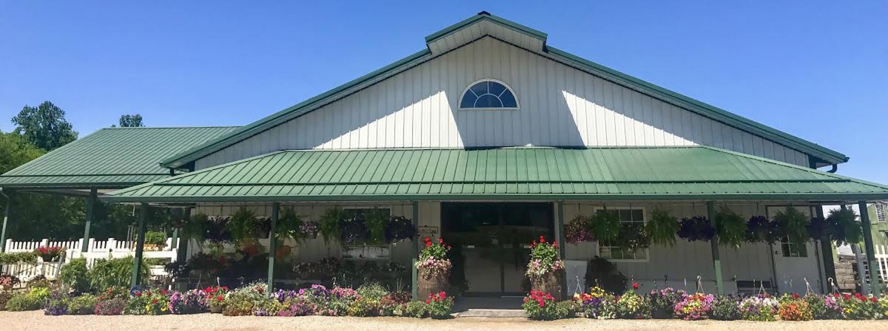 The white farm market with a green metal roof, hanging baskets and barrels of flowers